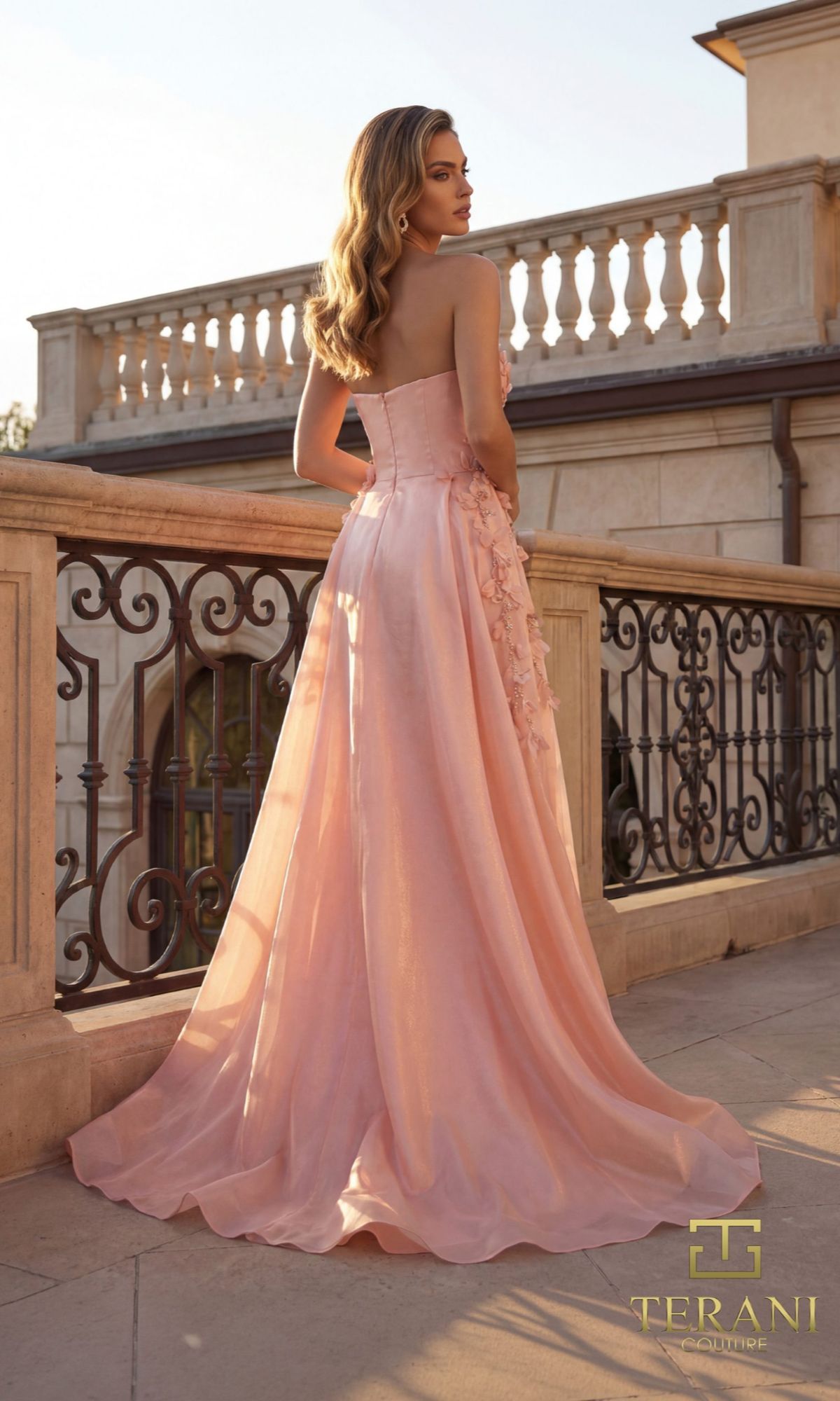 A woman in the Long Formal Dress: Terani 261E6195 stands on an outdoor terrace with ornate railings, facing away from the camera.